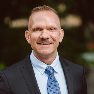 Headshot of Ricky Bradley in a black coat and blue shirt with a tie.