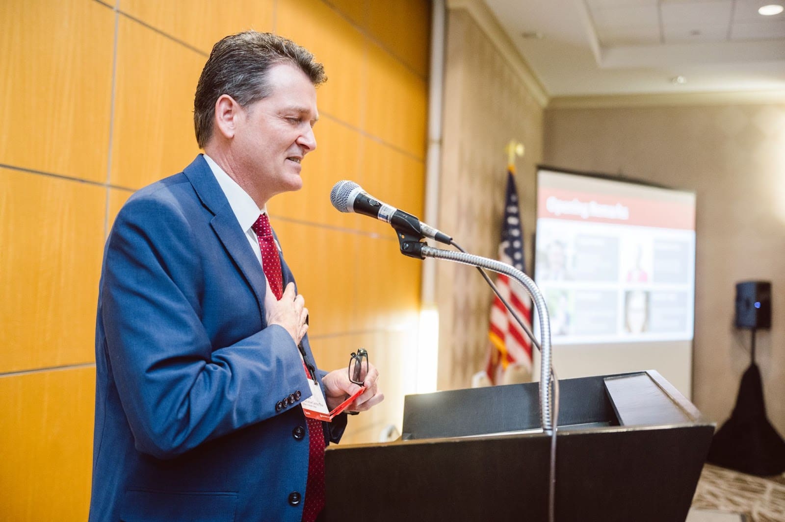 Man in blue suit and red tie stands at podium with microphone. His hand is over his heart.