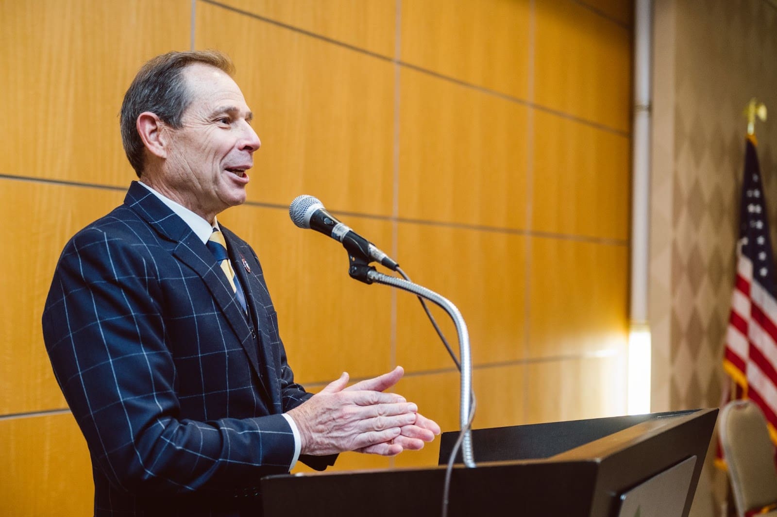Man in a checkered suit stands at a podium, speaking into a microphone. His hands are pressed together as if clapping.