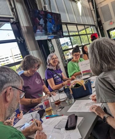 A candid photo shows people writing postcards at a restaurant table with big windows and a TV visible in the background