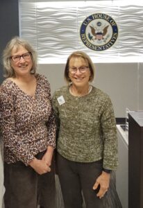 Two middle aged white women stand smiling in front of a wall showing the seal of the U.S. House of Representatives