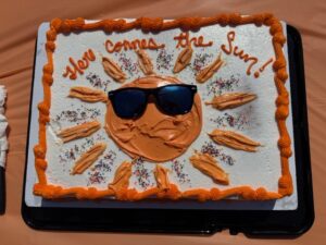 A cake decorated with an orange sun wearing sunglasses and the words Here Comes the Sun!