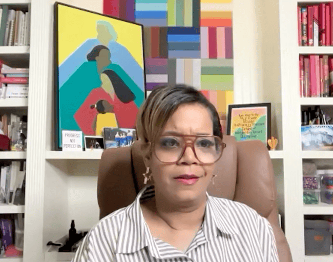 A Black woman wearing glasses and a collared shirt sits in front of colorful art in a home office.