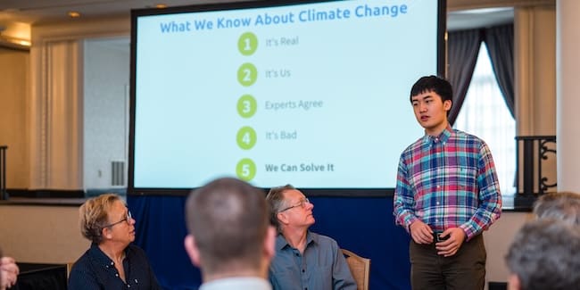 A young Asian man stands in front of a projector screen as if giving a presentation.