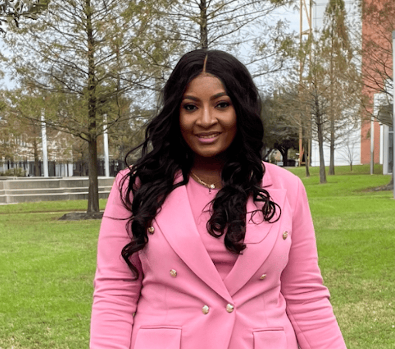 A brown-skinned woman with long black hair wears a pink suit.